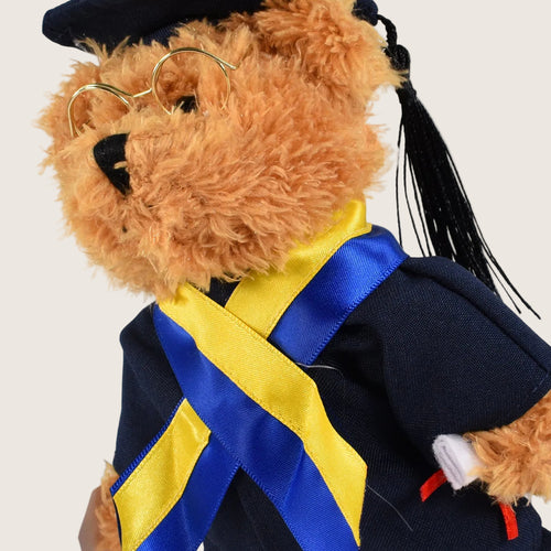 A plush teddy bear wearing a graduation outfit, including a navy blue gown, bright yellow and blue satin sash, gold-rimmed glasses, and a black cap with a tassel, shown against a plain white background.