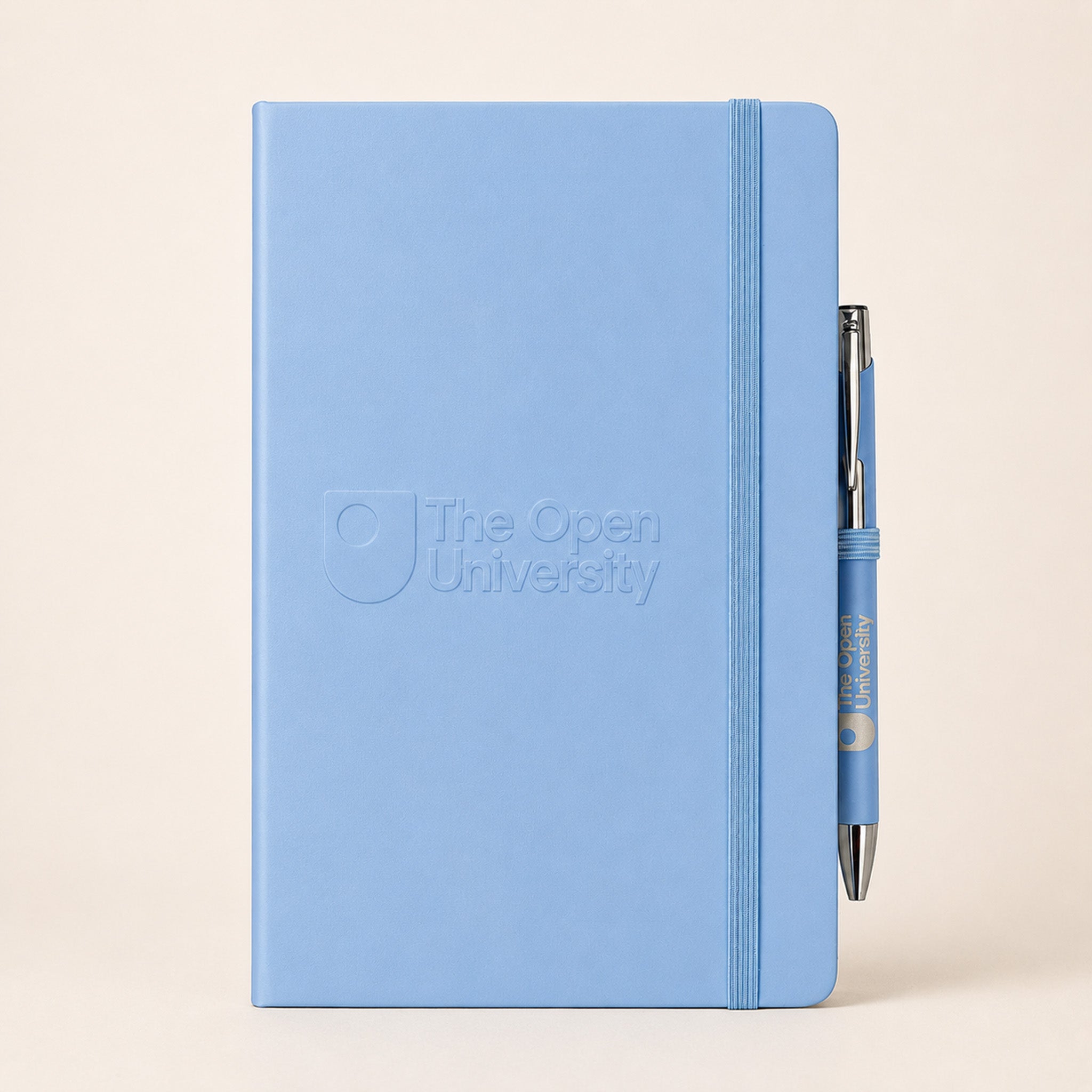 A light blue hardcover notebook with an embossed logo reading 'The Open University' on the front, shown standing upright with a matching blue pen tucked into the elastic closure against a warm beige background.
