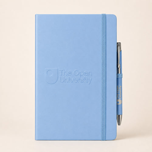 A light blue hardcover notebook with an embossed logo reading 'The Open University' on the front, shown standing upright with a matching blue pen tucked into the elastic closure against a warm beige background.