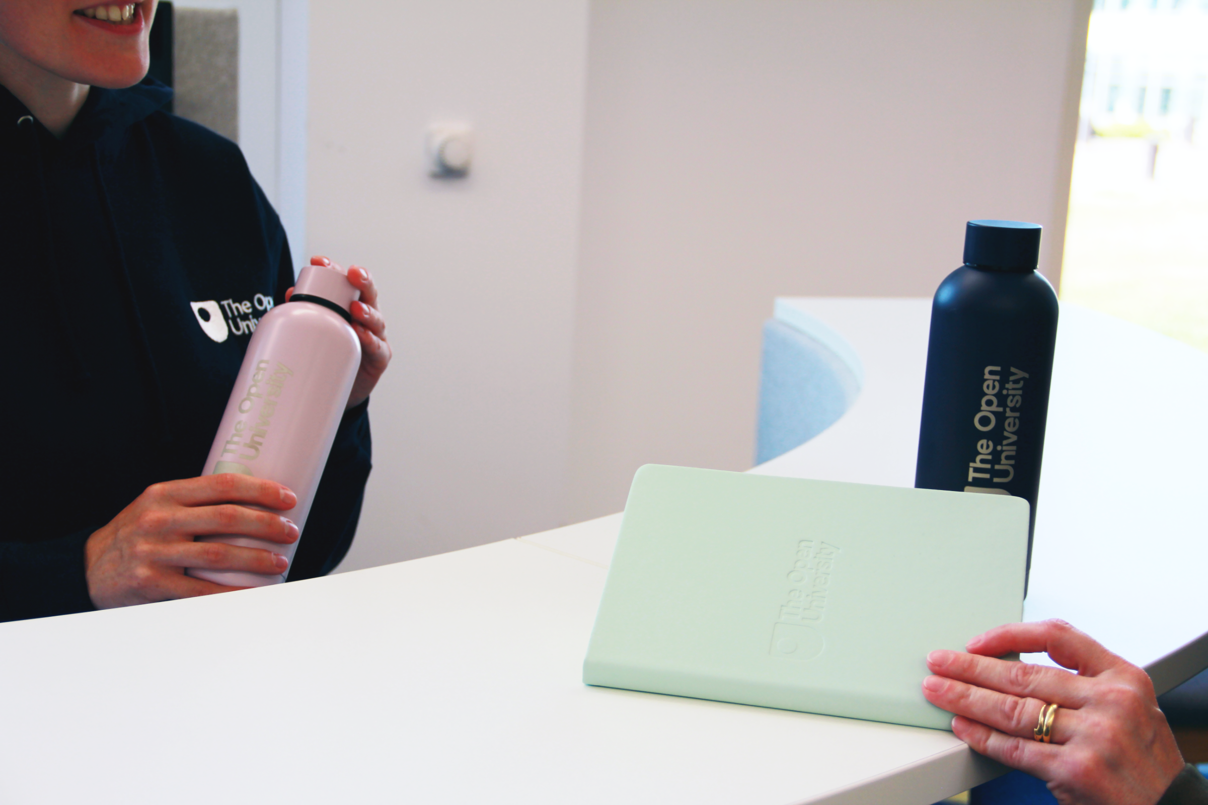 Two people holding branded water bottles at a white counter, with a mint green tablet in the foreground. One bottle is light pink with white text, the other is dark navy blue, and both read 'The Open University'.