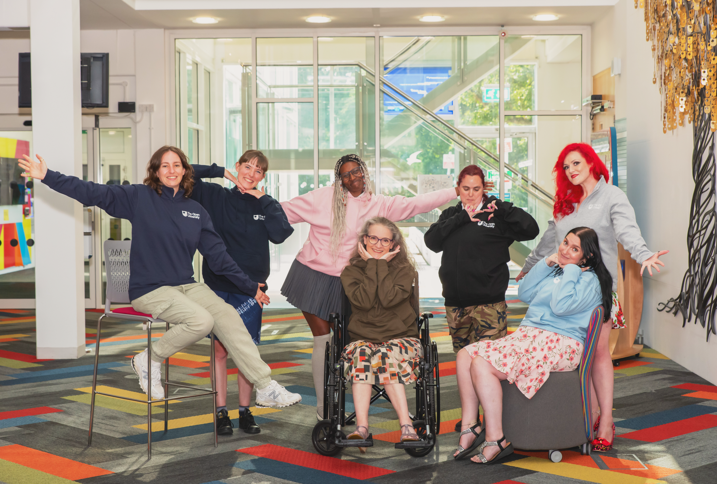 Six women posing together in a brightly lit modern indoor space with large windows, colorful carpet, and wall art. Several are wearing matching navy blue sweatshirts with the word 'VINTA' visible on the chest.