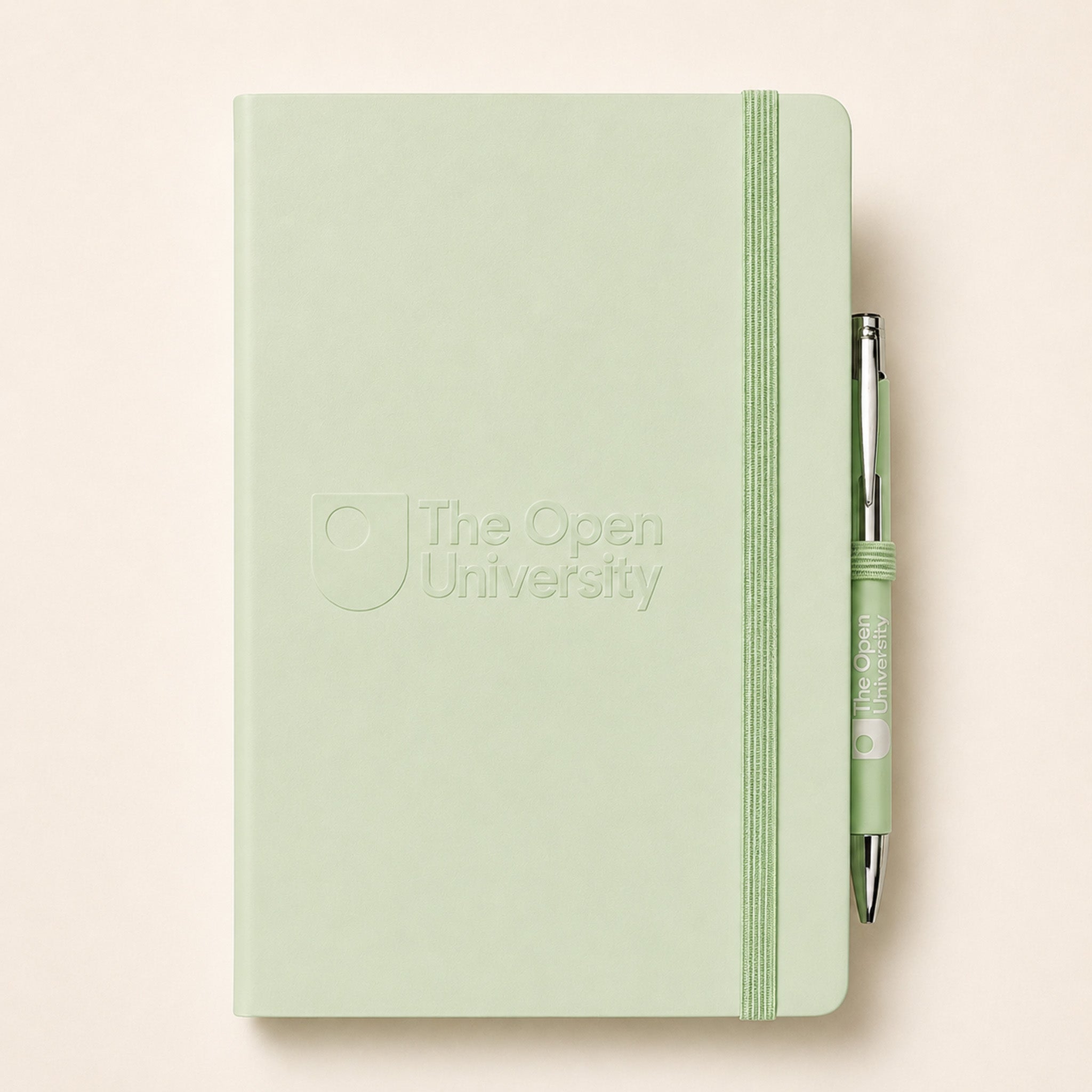A light sage green hardcover notebook with an elastic closure band and debossed 'The Open University' logo, shown with a matching green pen on a pale peach background.
