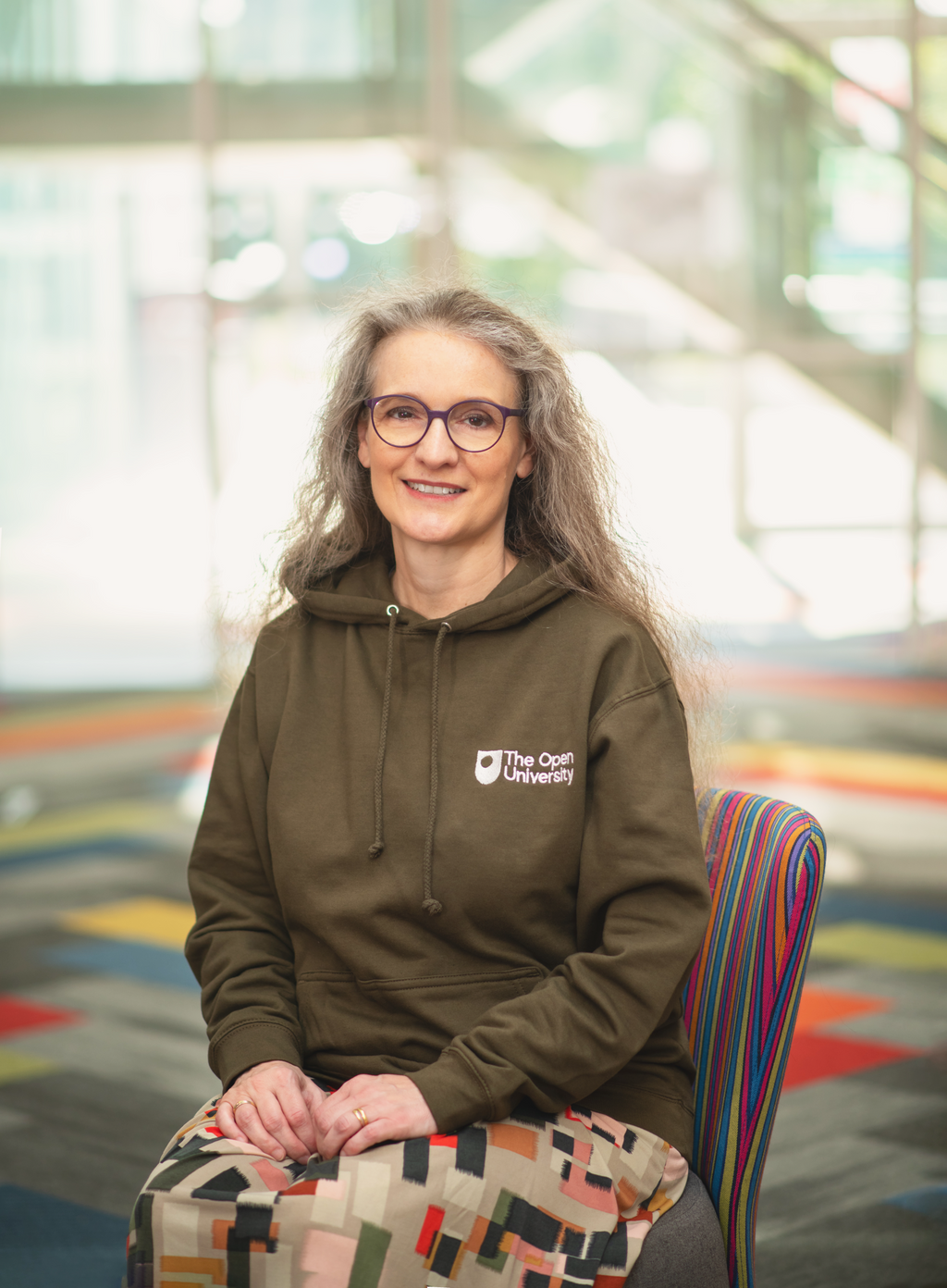 A woman with long gray hair and glasses sits smiling in a modern indoor setting, wearing an olive green hoodie with a white 'The Oet Van De University' logo on the chest and a multicolor geometric skirt.