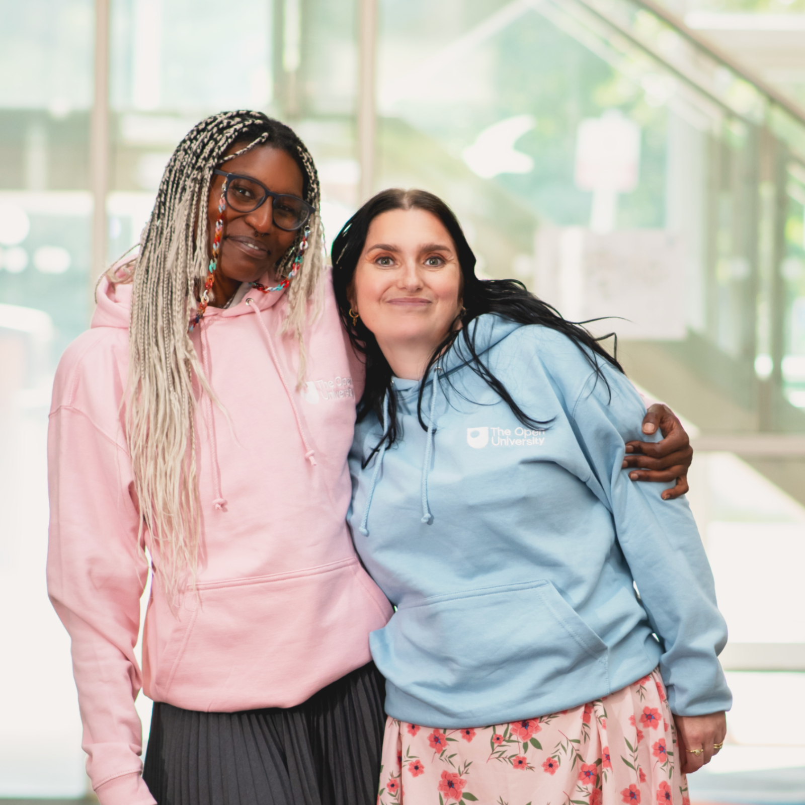 Two women standing together in a bright modern indoor setting, wearing matching branded hoodies. The woman on the left wears a pink hoodie, and the woman on the right wears a light blue hoodie with a white logo that reads 'The People's Trust Company'.