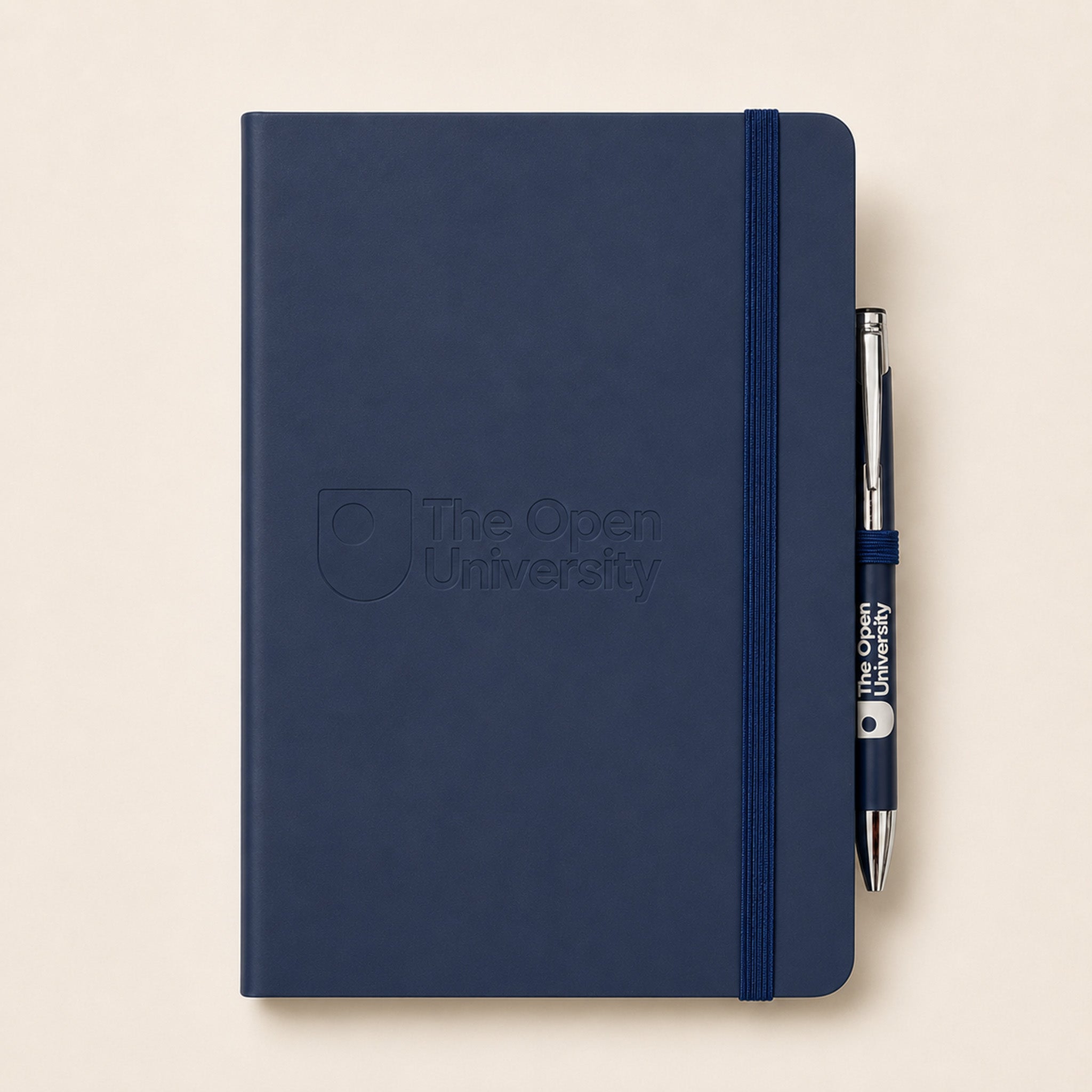 A navy blue hardcover notebook with an elastic closure band and an embossed 'The Open University' logo on the front, shown with a matching navy and silver pen on a light beige background.