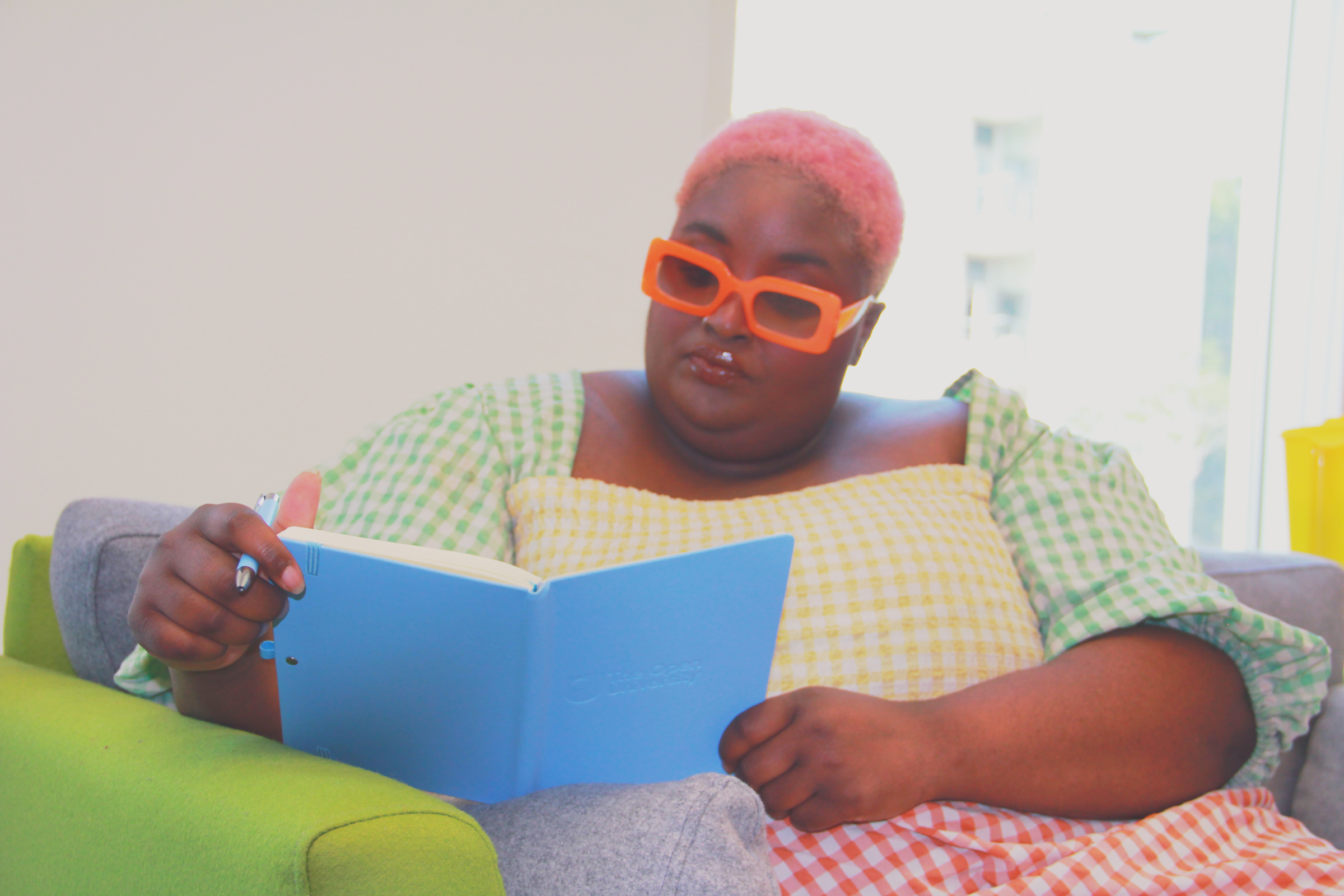 A person with pink hair and orange sunglasses sits on a couch reading a blue book, wearing a green and white gingham top and a yellow-and-white checkered pillow.