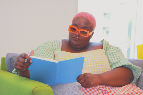A person with pink hair and orange sunglasses sits on a couch reading a blue book, wearing a green and white gingham top and a yellow-and-white checkered pillow.