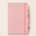 A pink hardcover notebook with an embossed 'The Open University' logo on the front, featuring a matching pink elastic closure band and a silver pen with a pink grip, set against a warm off-white background.