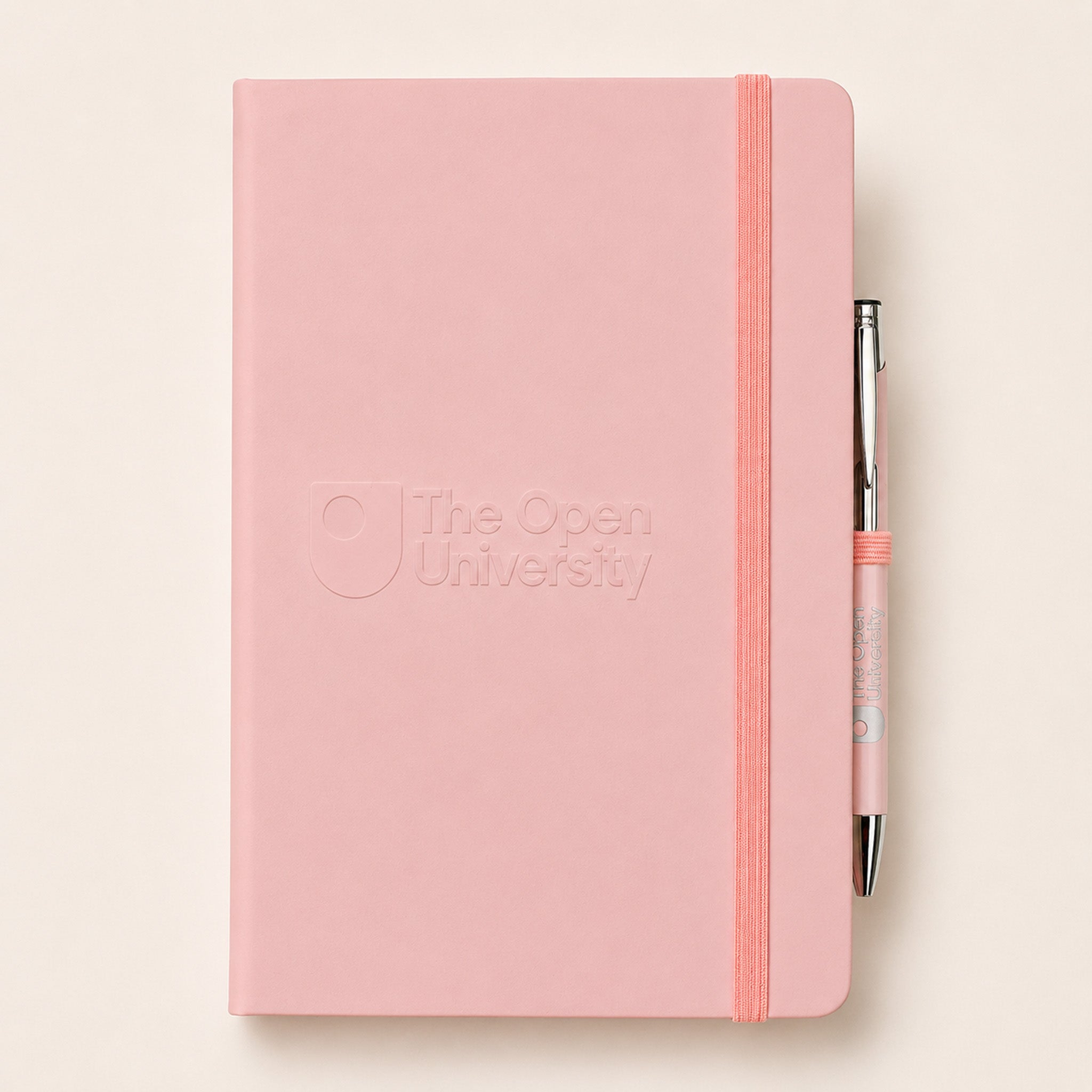 A pink hardcover notebook with an embossed 'The Open University' logo on the front, featuring a matching pink elastic closure band and a silver pen with a pink grip, set against a warm off-white background.