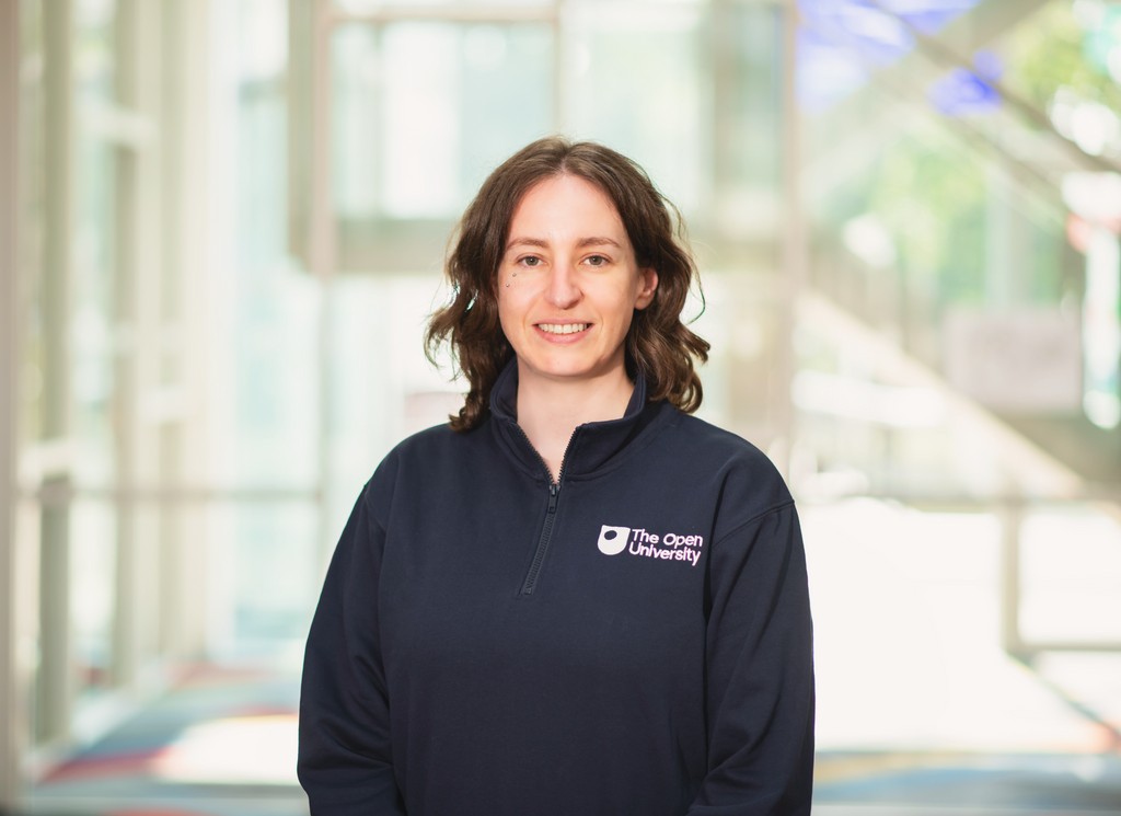 A woman with shoulder-length brown hair smiling, wearing a navy blue quarter-zip fleece pullover with a white 'The Open University' logo on the left chest, set against a bright, blurred modern building background.