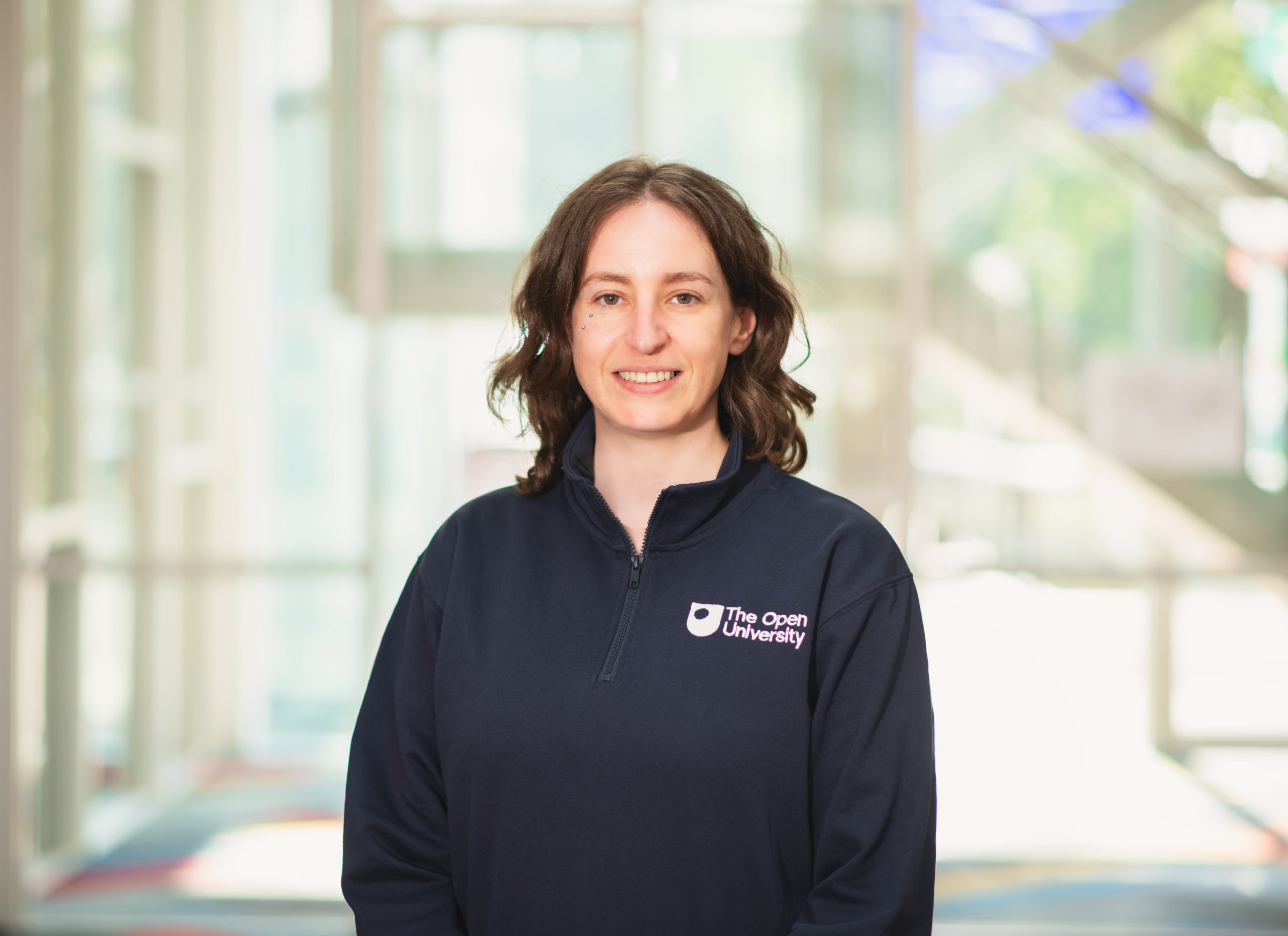A woman with shoulder-length brown hair smiling, wearing a navy blue quarter-zip fleece pullover with a white 'The Open University' logo on the left chest, set against a bright, blurred modern building background.
