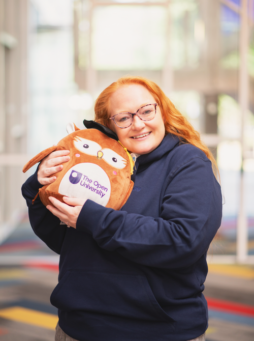 A smiling woman with red hair and glasses, wearing a navy blue hoodie, holds a brown plush owl toy with 'The Open University' logo on the front. The background is a brightly lit, blurred indoor space.