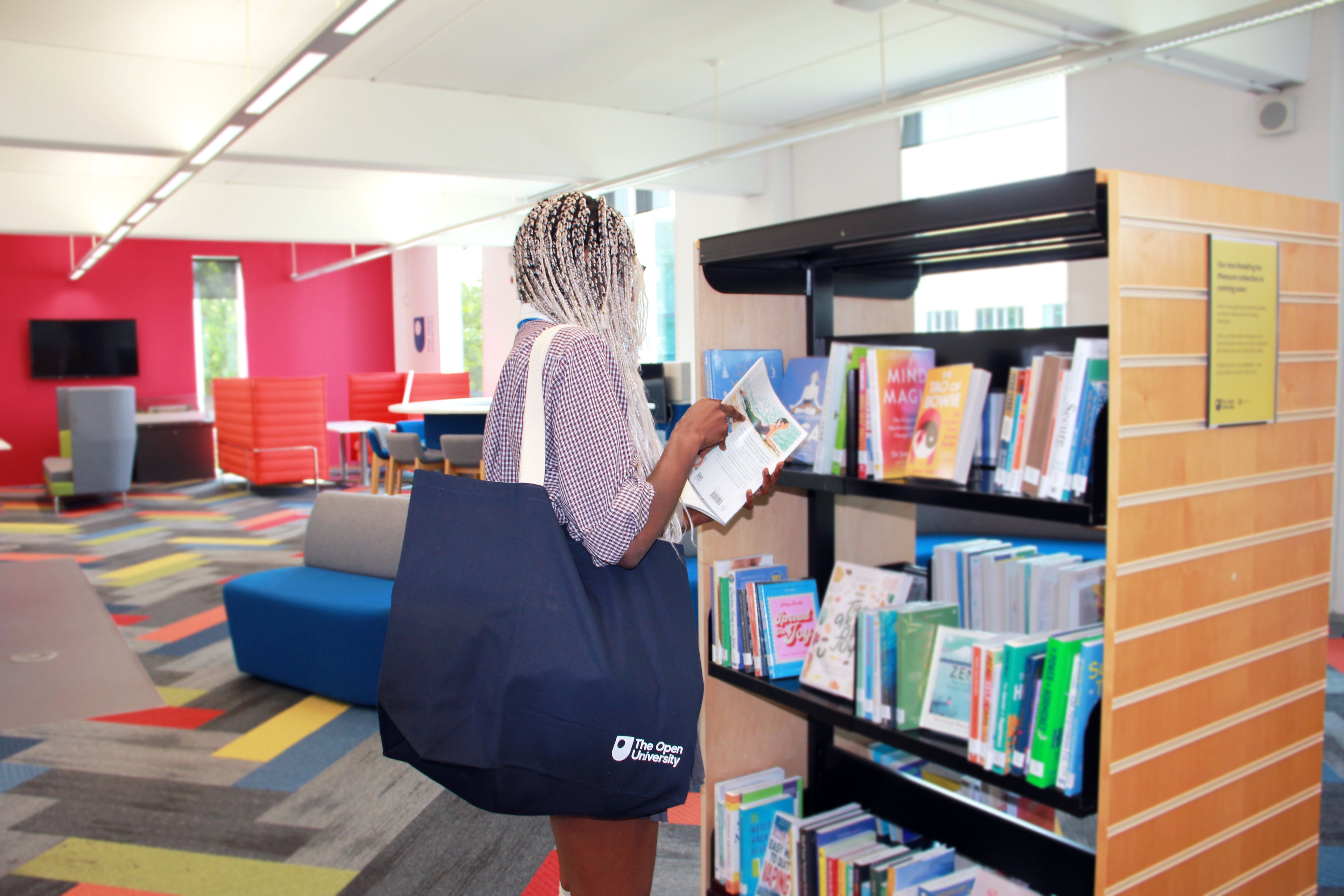 A woman in a library or bookstore stands in front of a bookshelf, holding a book and carrying a large navy tote bag with a white logo. The bag has 'The Print Society' printed on the front.